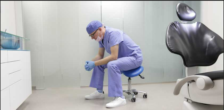 A dentist in blue scrubs sitting in a dental clinic beside the treatment chair